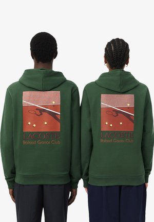 Due persone indossano felpe con cappuccio verdi con grafica di un campo da tennis, palline da tennis e la scritta "Lacoste Roland Garros Club" sul retro.