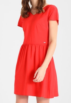Femme portant une robe rouge à manches courtes, longueur genou, avec une taille cintrée et un décolleté en V, debout devant un fond clair uni.