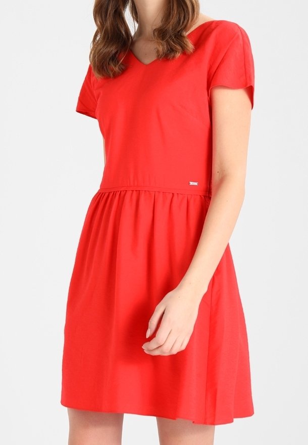Femme portant une robe rouge à manches courtes, longueur genou, avec une taille cintrée et un décolleté en V, debout devant un fond clair uni.