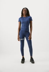 Kvinne står i marineblå Nike treningsantrekk, inkludert kortermet topp og leggings med logoer, iført svarte joggesko, på hvit bakgrunn.