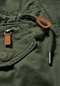 Olivově zelená látka s texturovaným povrchem, kovovým zipem a koženou označovací visačkou připevněnou k detailu se šňůrkou.