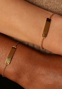 Goudkleurige armbanden met rechthoekige metalen platen en delicate schakels, weergegeven op lichte en donkere huidtinten. Gladde afwerking.