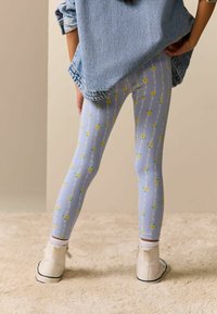 Modré legíny s květinovým vzorem žlutých květin, nošené s džínovou bundou a bílými kotníkovými botami.