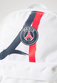 Mochila blanca con un diseño abstracto en negro, azul y rojo. El logo del Paris Saint-Germain está prominently exhibited. Material resistente con detalles de costura.