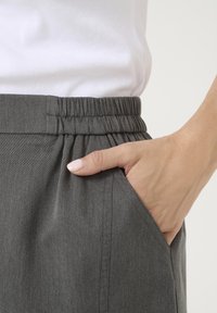 Pantaloni grigi leggeri con vita elastica arricciata, dotati di una tasca laterale e texture liscia. Abbinati a un top bianco aderente.