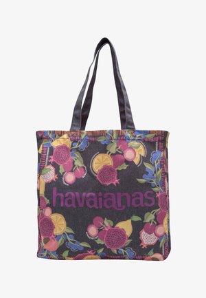 Bolsa tote con fondo oscuro, patrones coloridos de frutas y logo morado de "havaianas", con dos asas largas y oscuras.