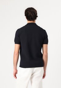 Marineblaues kurzärmliges Strick-Polohemd mit geripptem Kragen und Saum. Verfügt über einen strukturierten Stoff und eine figurbetonte Passform. Getragen mit hellen Hosen.