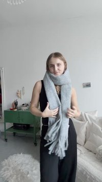 Grå skjerf laget av mykt materiale, med frynsete ender. Bæres løst rundt halsen, kombinert med et svart antrekk i et lyst miljø.