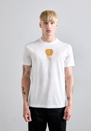 Noormees, kellel on blondid juuksed, kannab valget Versace T-särki, millel on kuldne Medusa logo ja tekst, musti pükse ning seisab ühetaolise heleda tausta ees.