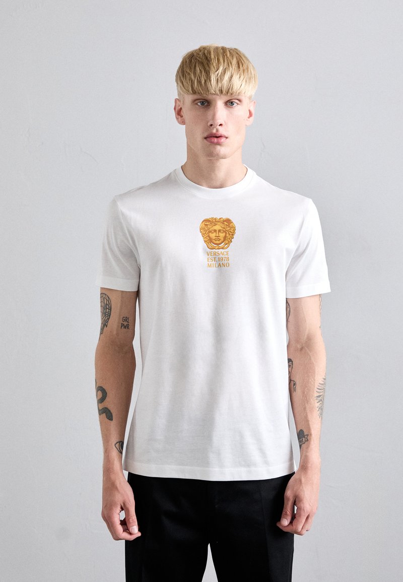 Young man with blonde hair wearing a white Versace t-shirt with gold Medusa logo and text, black pants, standing against a plain light background.