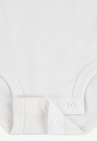 Bodystocking kratkih rukava, boje krem, izrađen od mekanog pamuka, s kopčama na gumbe i glatkom, blago teksturiranom tkaninom.