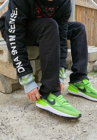 Gröna Nike-sneakers med en strukturerad ovandel, svarta accenter och vita sulor. En person knyter sina skor medan hen har på sig mörka byxor och en svart sweatshirt.