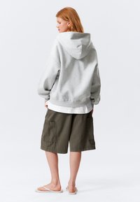 Grå hettegenser med senket skuldre, ribbestrikket kant, og sidespalter, kombinert med olivengrønne cargo-shorts og beige sandaler.