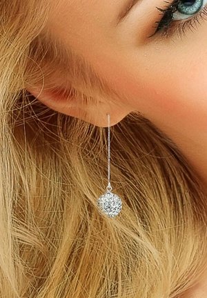 Les boucles d'oreilles en chaîne argentée présentent un pendentif rond en rhinestone, pétillant avec diverses facettes sur fond de cheveux blonds clairs.