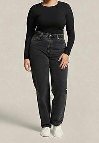 Personne portant un haut noir moulant à manches longues, un jean noir taille haute et des baskets blanches, debout devant un fond uni.