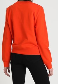 Oranžová mikina s kulatým výstřihem, žebrovanými manžetami a lemem. Hladký materiál s volnějším střihem, kombinovaná s černými kalhotami.