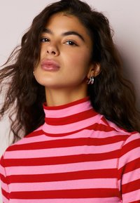 Rolákový top s červenými a ružovými pruhmi a rebrovanou textúrou, s priliehavým strihom a vysokým golierom. Vlasy upravené do voľných vĺn.