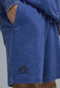 Pantaloni corti di cotone blu con una texture scolorita, dotati di tasche laterali e un logo nero di Sonic the Hedgehog sulla gamba sinistra.