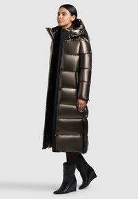 Langer Puffer-Mantel in metallischem Bronze, ausgestattet mit einer Kapuze, großen gesteppte Abschnitten und seitlichen Reißverschlusstaschen. Kombiniert mit schwarzen Wedge-Ankle-Boots.