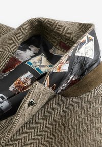 Veste marron à motif chevrons avec une doublure intérieure noire présentant des motifs colorés de timbres-poste.