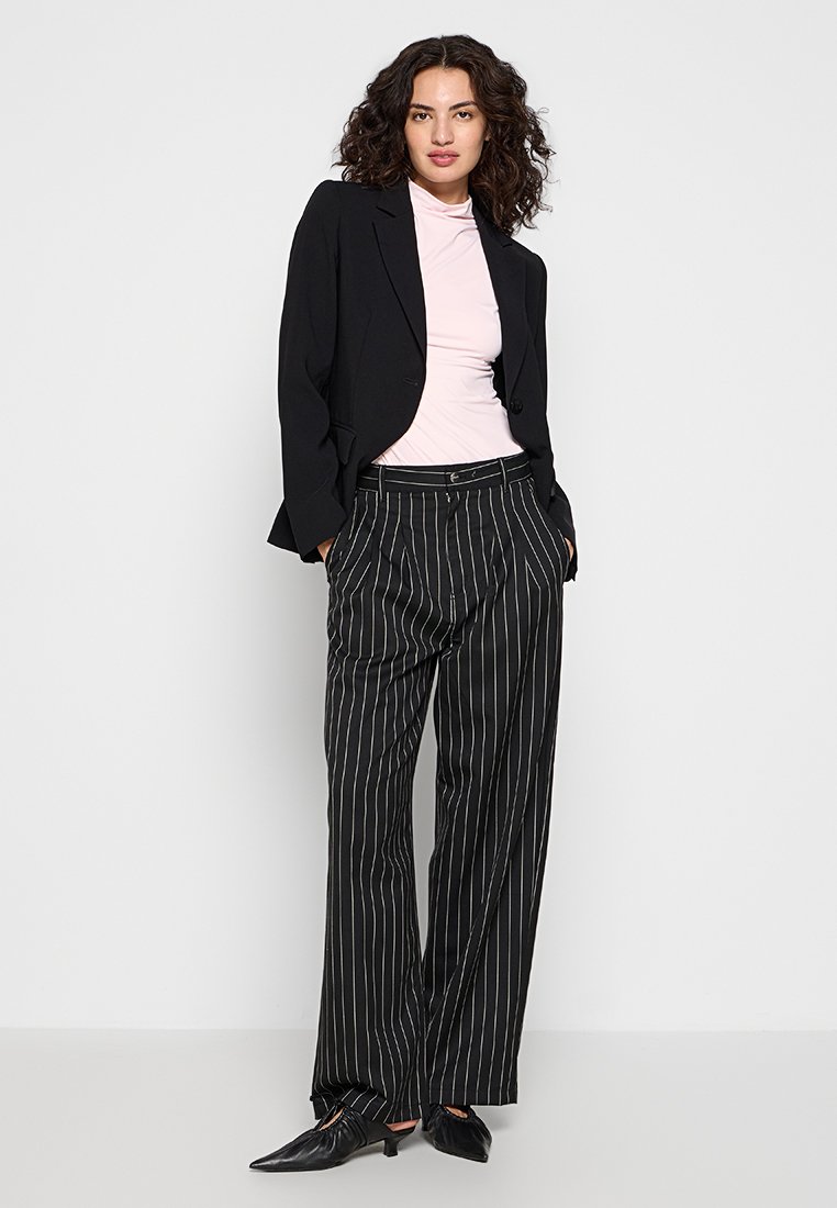 Blazer noir sur un haut rose à col montant, associé à un pantalon large à rayures noires et blanches. Ballerines noires à bout pointu. Textures lisses et coupe ajustée.