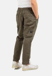 Person wearing olive green cargo pants and white sneakers, standing with hands in pockets, shown from mid-back to feet.