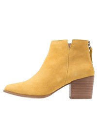 Botas de ante amarillas hasta el tobillo con puntera en punta, cremallera lateral y tacón de madera apilada. Textura suave con detalles de costura mínimos.