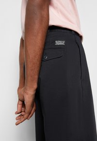 Pantalones negros de tejido suave, con un bolsillo trasero con botón y etiqueta de la marca. Mano descansando en el lado, mostrando el ajuste y el diseño.