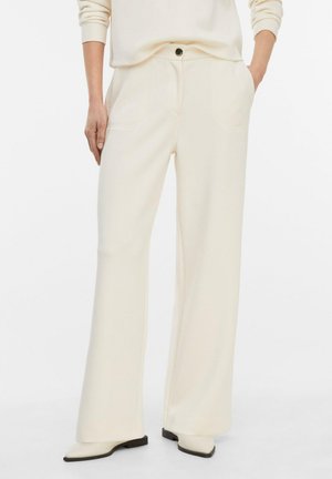 Kobieta ubrana w szerokie spodnie w kolorze ecru z przednimi kieszeniami i czarnym guzikiem, w połączeniu z dopasowanym długim rękawem i białymi butami na spiczasty nosek.