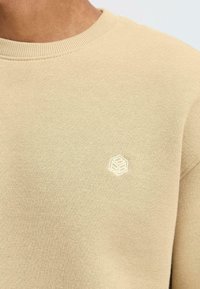 Beige sweatshirt with ribbed crew neck. Features a small embossed geometric logo on the chest. Soft, textured fabric.