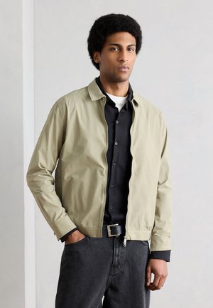 Mand med kort krøllet hår iført beige lynlåstrøje over en sort skjorte, sort bælte og mørkegrå jeans, stående mod en lys baggrund.