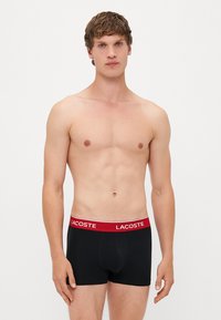 Boxer shorts noirs avec une ceinture élastique rouge portant le nom de la marque "Lacoste" en blanc. Le tissu semble lisse et ajusté.