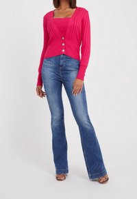 Kobieta w jaskraworóżowym, prążkowanym kardiganie zapinanym na guziki, noszącym dopasowaną bluzkę, w połączeniu z wyblakłymi, niebieskimi spodniami typu bootcut i sandałami na obcasach z odkrytymi palcami.