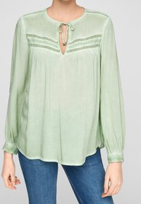 Blusa verde chiaro con maniche lunghe, presenta un collo rotondo, dettaglio con fiocco e cuciture decorative sul petto. Tessuto morbido.