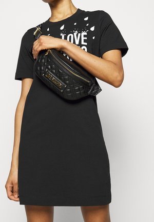 Marsupio nero trapuntato con una placca logo dorata e chiusura a zip, indossato a tracolla. Il modello indossa un semplice vestito t-shirt nero.