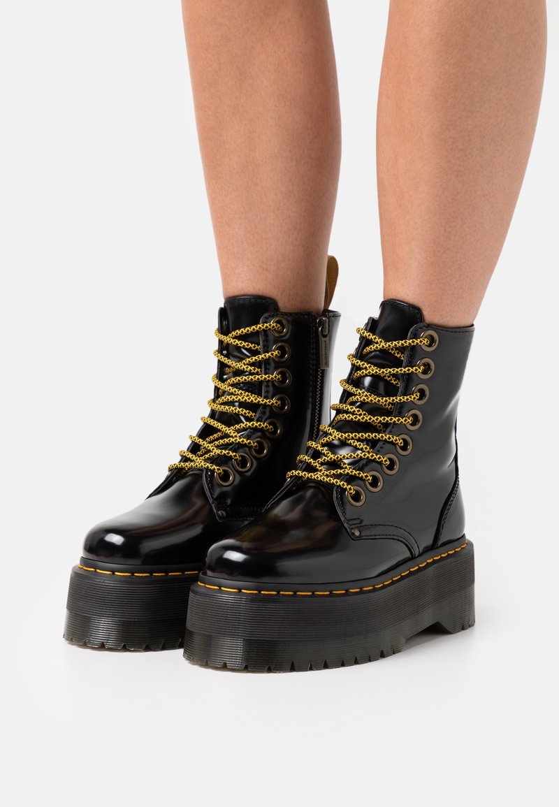 Bottes à plateforme noires brillantes avec semelles épaisses, lacets jaunes et noirs, et fermeture éclair latérale portées sur des jambes nues sur fond blanc.