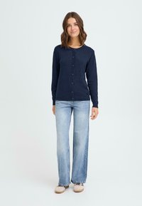 Marineblå cardigan med knapfront, lange ærmer og rund halsudskæring, parret med lyseblå flagrende jeans og afslappede beige sko.