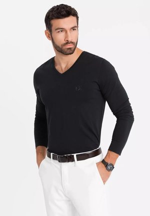 Camisa negra de manga larga con escote en V, hecha de un tejido suave, combinada con pantalones blancos y un cinturón marrón. Un sutil logo aparece en el pecho.