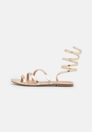 Gyldne metaliske flade sandaler med fire horisontale stropper og en spiralformet ankelstrop. Glat tekstur, minimalistisk design, åben tå-stil.