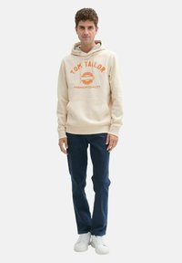 Beige Kapuzenpullover mit orangem Druck, frontale Kängurutasche, Rippbündchen, kombiniert mit dunkelblauen Jeans und weißen Sneakers.