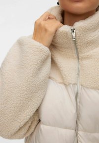 Person iført en cremefarvet sherpa fleecejakke med høj krave, der holder kraven nær den metal lynlås.