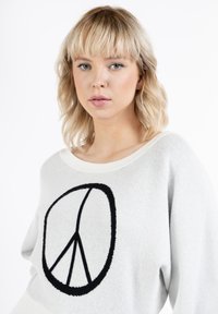 Maglione grigio chiaro con grafica del simbolo della pace nera. Il tessuto appare morbido, con una vestibilità comoda e un ampio scollo.