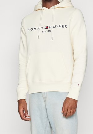 Man wearing cream hoodie with "Tommy Hilfiger EST. 1985" text and logo on chest and sleeve, paired with light blue jeans.