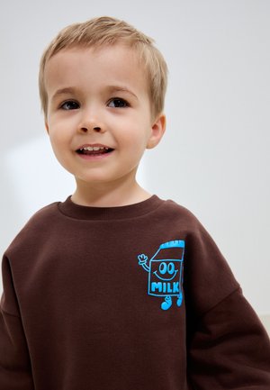 Ung dreng smilende, iført en brun sweatshirt med en blå tegneserie mælkekarton, der vinker på brystet.