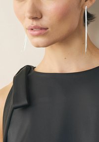 Sølvøreringe med hængende, slankt design hænger ved siden af en sort, ærmeløs kjole med draperet skulderdetalje.