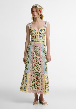 Nő virágmintás hímzett crop topot és hozzáillő, pasztell panelekkel díszített midi szoknyát visel, melyhez virágos fejkendő és bézs ék sarkú szandál társul.