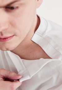 Biała bawełniana koszula z zapięciem na rzep przy kołnierzyku. Materiał jest gładki, a projekt zawiera widoczną linię dekoltu.