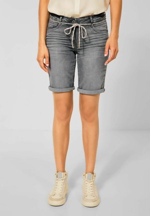 Mujer con shorts de mezclilla grises remangados con cintura de cordón y zapatillas beige de caña alta sobre fondo gris.
