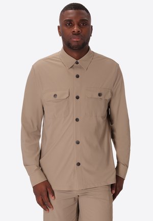 Uomo che indossa una camicia beige con bottoni e due tasche sul petto e pantaloni beige abbinati su sfondo semplice.