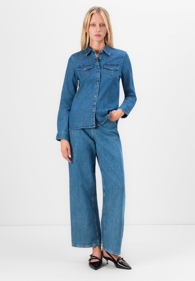 Camisa de mezclilla clara con cierre de botones y dos bolsillos en el pecho, combinada con pantalones de mezclilla de pierna ancha. Ambos son de un tono azul uniforme.